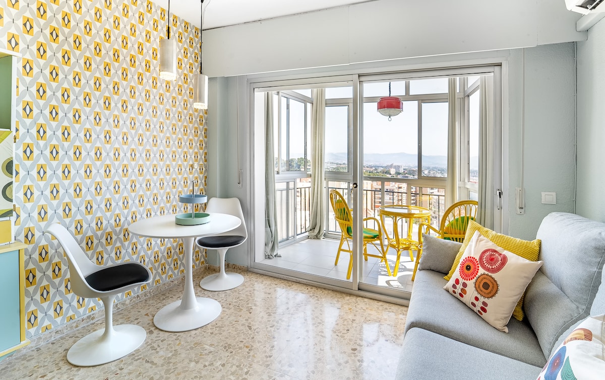 A bright living area features a soft gray sofa adorned with colorful cushions. A white round table is accompanied by two contrasting chairs, creating an inviting dining space. Large glass doors open to a balcony with scenic views, while patterned wallpaper adds a playful touch.