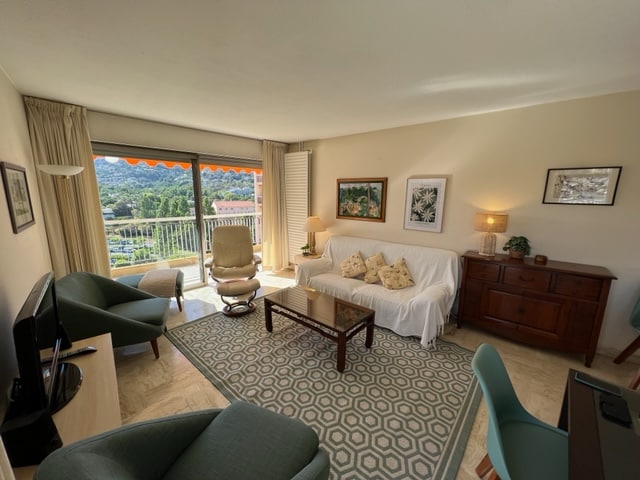 A bright living room features large windows opening to a scenic view. A comfortable sofa, accent chairs, and a wooden coffee table create a welcoming space. Decorative artwork adorns the walls, and a sideboard provides storage. Natural light enhances the warm ambiance of the room.