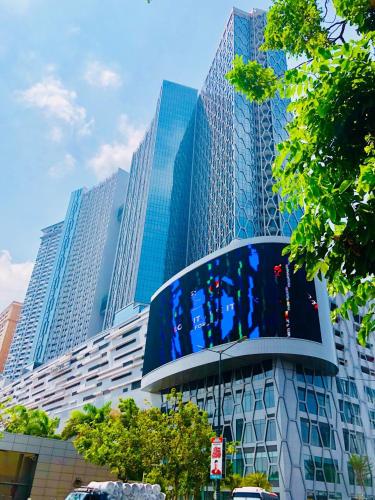 A modern, multi-story building showcases a unique architectural design with a combination of glass and metal surfaces. Lush greenery surrounds the entrance, creating a contrast with the sleek structure. A large digital screen is prominently displayed at the base of the building.