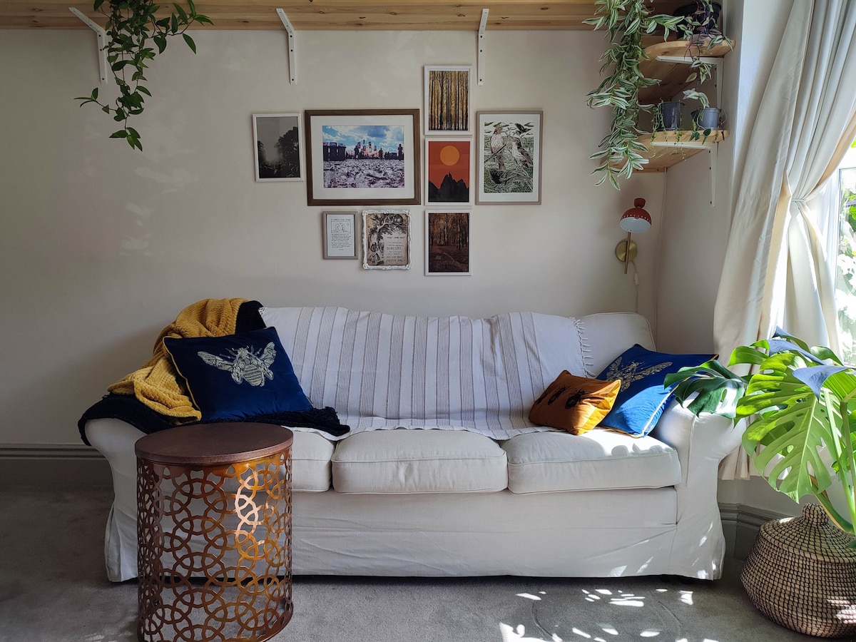 A white sofa with a striped throw is positioned against a light wall adorned with framed photographs and artwork. Decorative cushions in deep blue and mustard complement the arrangement. A small, patterned side table and lush greenery enhance the inviting, cozy ambiance.
