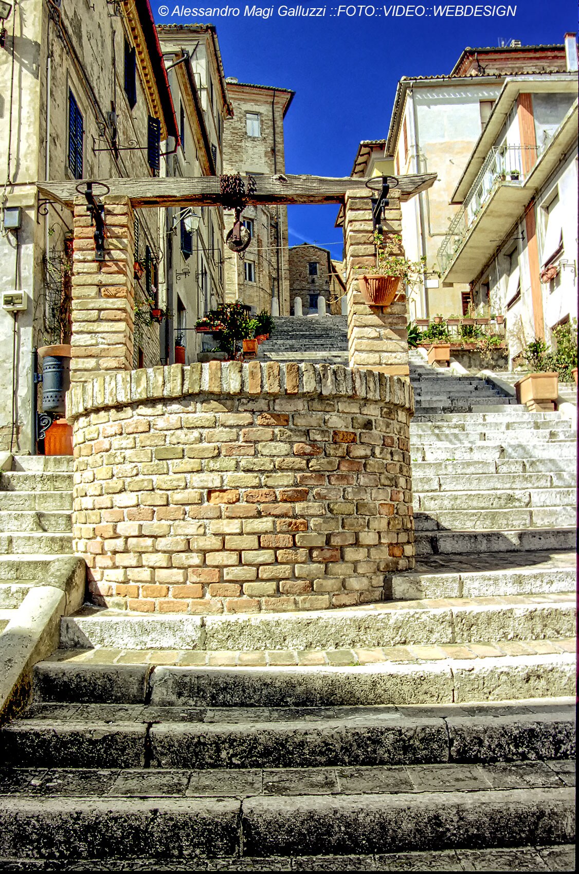 A charming stone well is positioned at the base of a staircase, surrounded by traditional buildings. The well features brickwork with hanging lanterns and flower pots. The steps lead upwards, framed by quaint architecture under a clear blue sky.