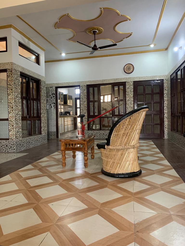 A spacious lobby area is presented, showcasing a decorative floor pattern of alternating wood and tile finishes. A traditional woven chair is positioned beside a rustic wooden table, with large windows allowing ample natural light. The room is framed with intricately designed walls and a ceiling fan overhead.