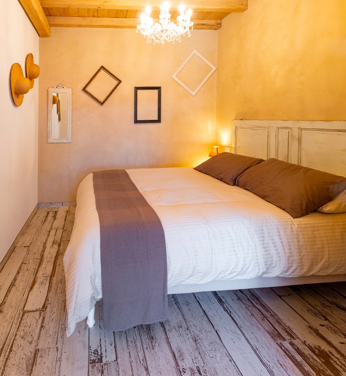 A cozy bedroom features a spacious bed with a neutral-toned duvet and two pillows. Soft lighting is provided by bedside lamps and a decorative chandelier. The wooden ceiling and rustic floor complement the warm wall tones, while framed mirrors and wall art add a touch of character.