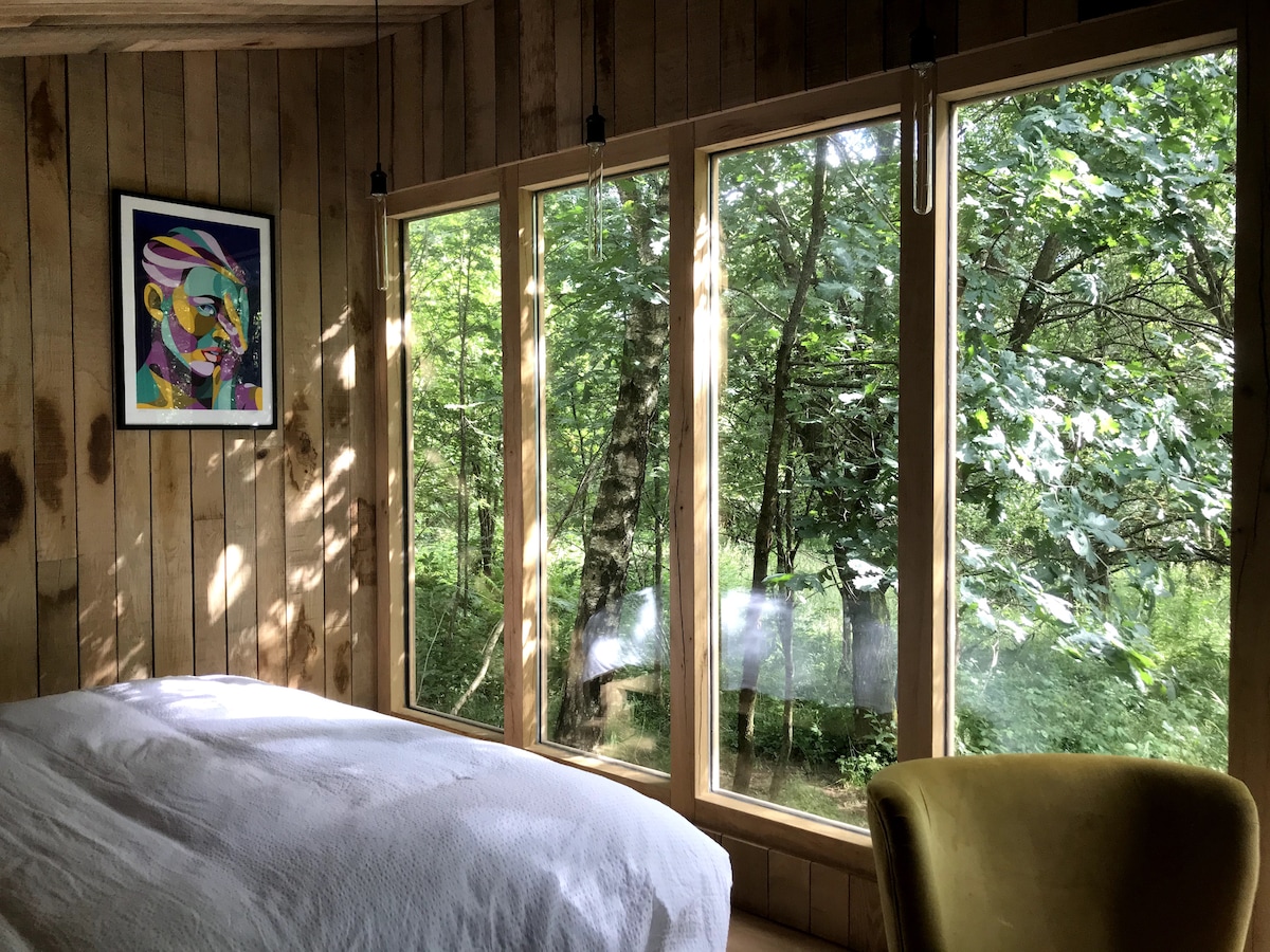 A cozy sleeping area features a spacious bed covered with a white comforter, complemented by a green accent chair. Large windows allow natural light to flood the room, offering a view of the surrounding trees and greenery, enhancing the peaceful atmosphere.