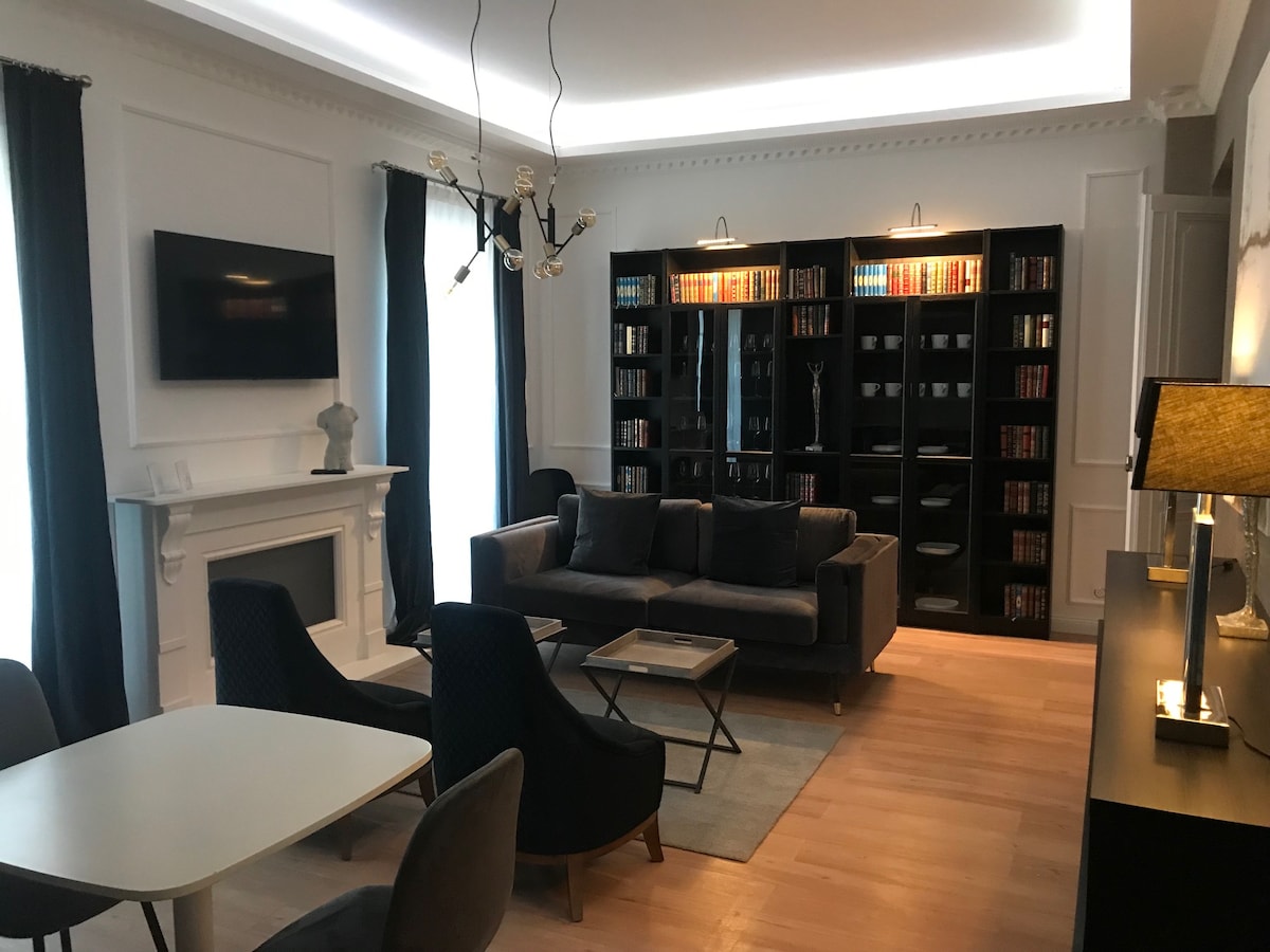 A stylish living room features a gray sofa with multiple dark cushions, positioned in front of a decorative fireplace. A bookshelf filled with books lines one wall, while an elegant light fixture hangs from the ceiling. Large windows are framed by classic dark curtains.