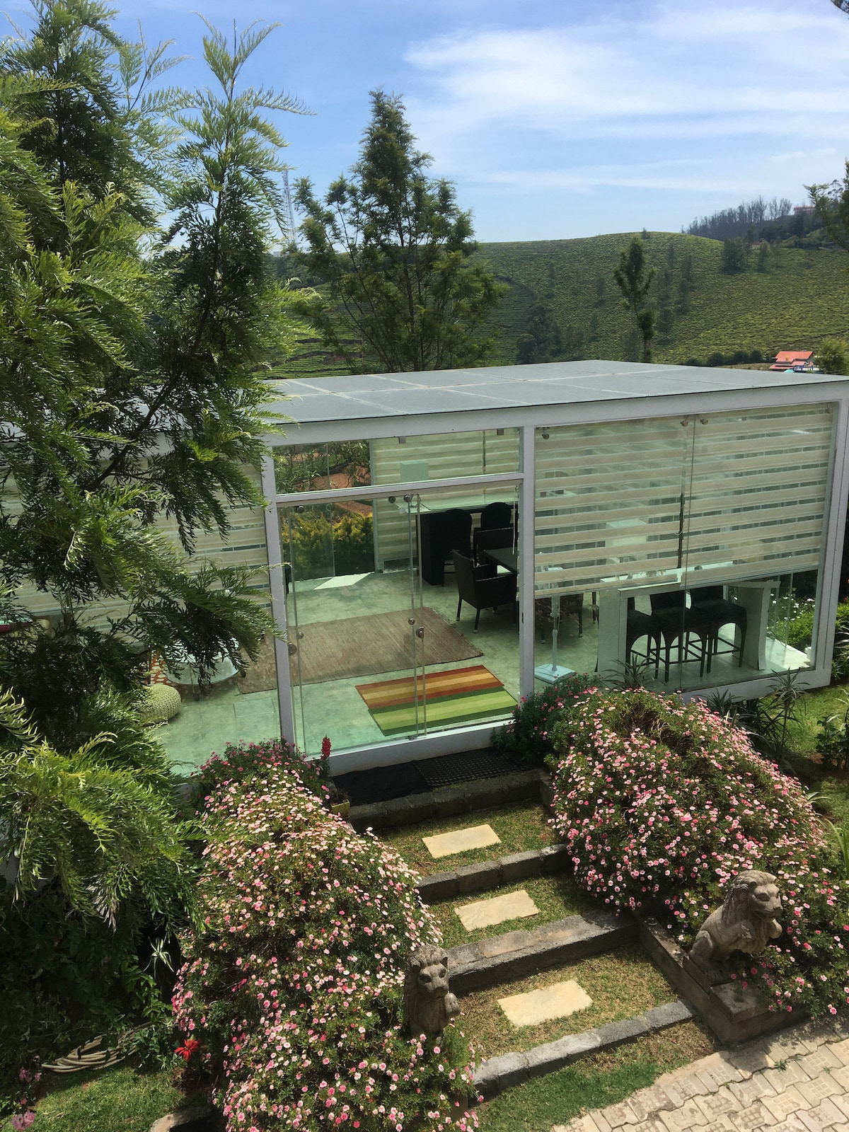 A modern glass structure is surrounded by lush greenery and blooming flowers. The transparent walls allow for natural light, revealing a cozy lounge area and dining space within. Stone pathways lead up to the entrance, enhancing the serene outdoor setting.