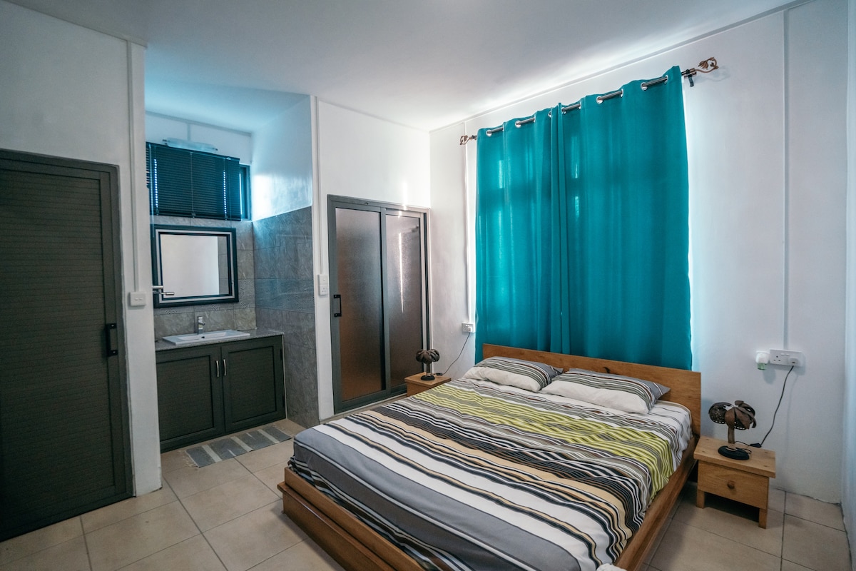 A bright bedroom features a spacious bed with a striped coverlet, positioned near large windows adorned with aqua curtains. A mirrored dresser and modern light fixtures complement the room's neutral tones, while an en-suite bathroom is visible in the background.