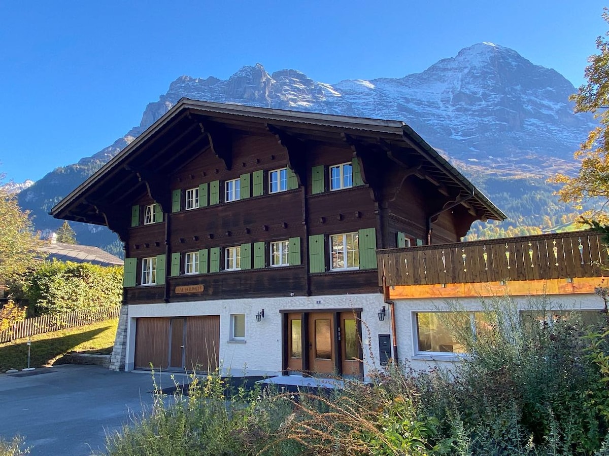 A traditional chalet-style building features a wooden exterior with green shutters, set against a backdrop of snow-capped mountains. Spacious parking is visible in front, and a balcony extends from the upper floor, offering additional outdoor space.