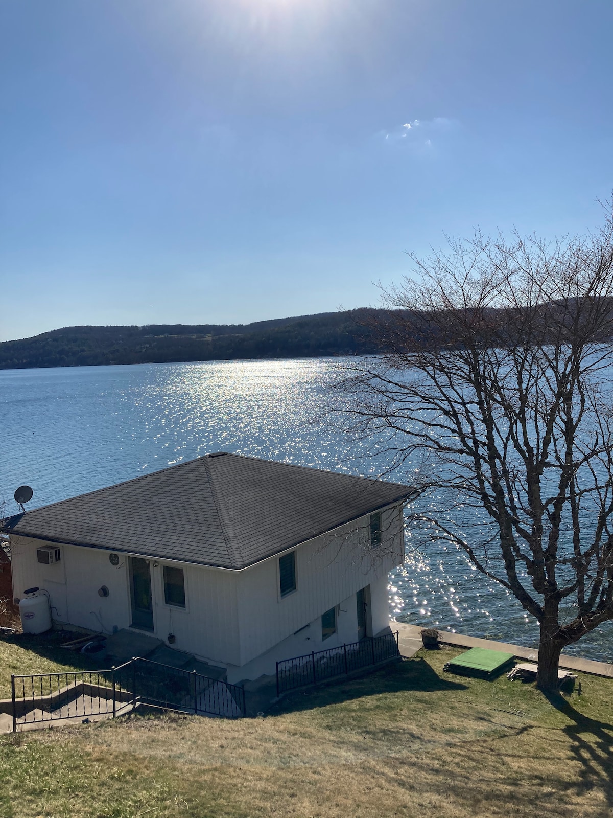 The exterior of a white boathouse is visible, situated on the shore of Otsego Lake. Sunlight glimmers on the water's surface, while a bare tree stands nearby. Gentle hills are seen in the background, adding to the serene lakeside setting.