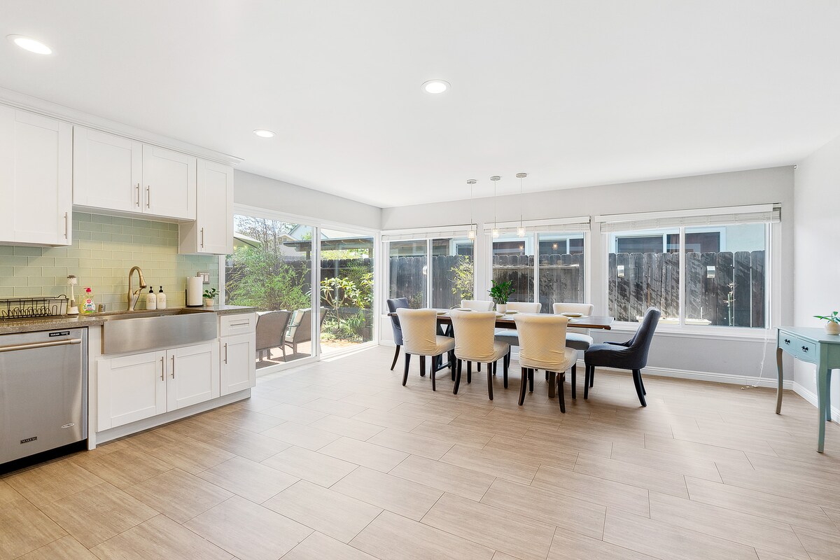 A bright and airy kitchen and dining area features modern white cabinetry and stainless steel appliances. Natural light flows through large windows, illuminating the spacious dining table with beige upholstered chairs. The kitchen countertop is complemented by a mint green backsplash and overlooks the greenery outside.