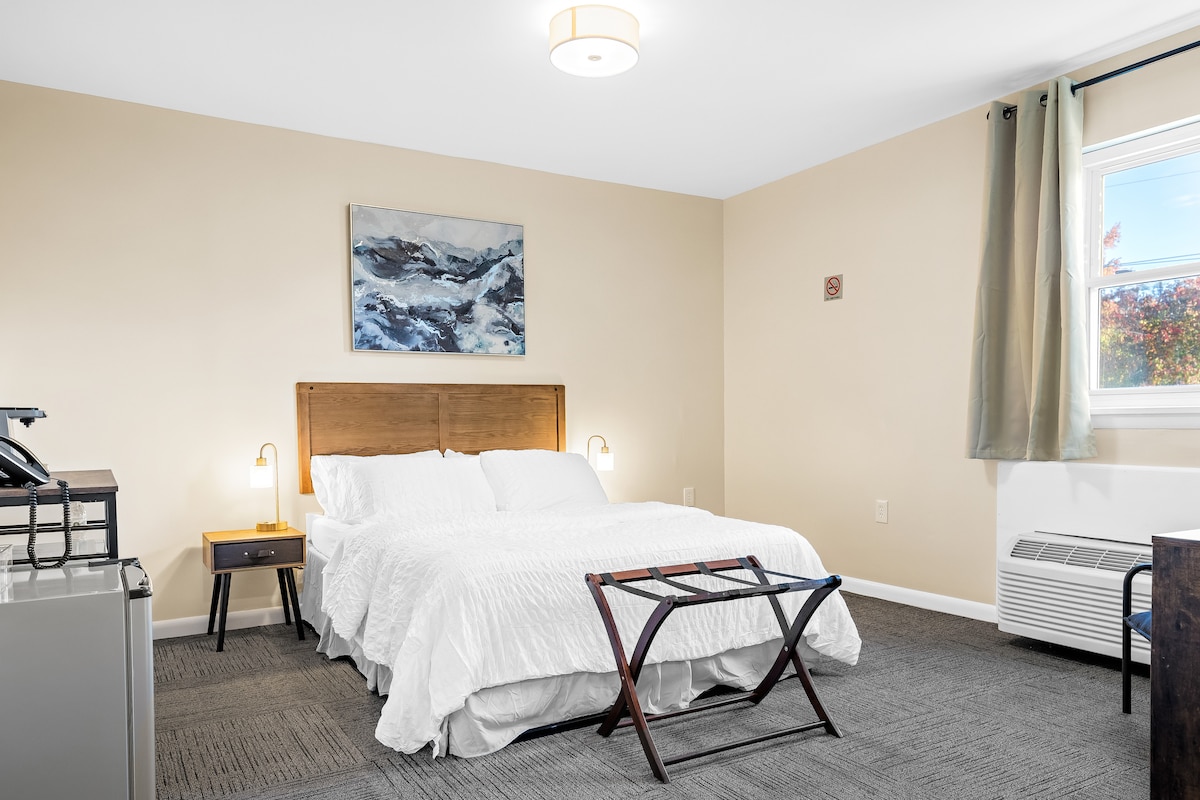The bedroom features a spacious bed with a light-colored quilt and wooden headboard. A small bedside table with a lamp is positioned beside the bed. Natural light enters through a window with light curtains, illuminating the neutral-toned walls and carpeted floor.