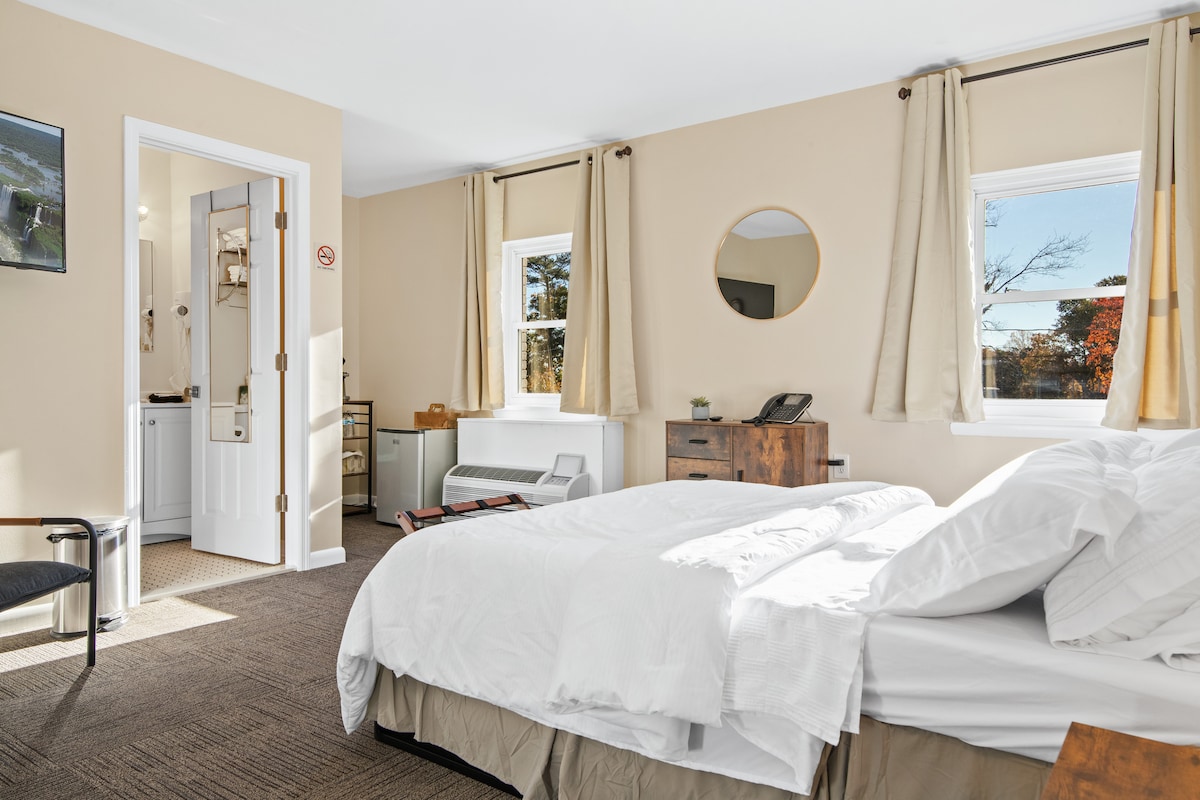 A well-lit room features a spacious bed with white linens, a wooden nightstand, and a mirror. Natural light enters through two windows with light curtains. A kitchenette with a mini refrigerator and microwave is visible, along with a chair and a door leading to the private bathroom.