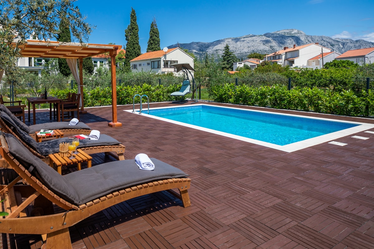 A private pool area is visible, featuring a clear blue pool surrounded by wooden decking. Lounge chairs are arranged nearby, with a table set for outdoor dining. Lush greenery lines the perimeter, and mountains provide a scenic backdrop under a clear blue sky.