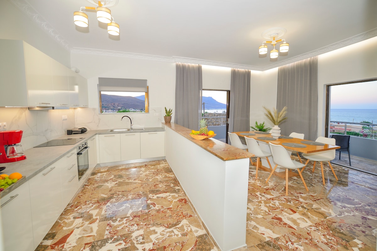 A modern kitchen and dining area are presented with light-colored cabinetry and ample counter space. Natural light floods in through large windows, revealing views of the sea. A dining table is complemented by white chairs, and a countertop is adorned with fresh fruit.
