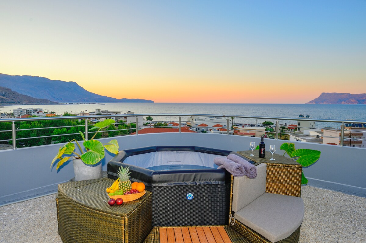 A private rooftop garden features a heated jacuzzi surrounded by lounge seating and tables. Fresh fruit is displayed on a wooden surface, with potted plants adding greenery. The panoramic view includes the sea and distant mountains during sunset, providing a serene atmosphere.