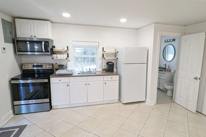The kitchen features modern appliances, including a stainless steel stove and microwave, a white refrigerator, and a coffee maker. Ample counter space is provided, accompanied by white cabinetry. A window allows natural light to brighten the work area, with a door leading to a bathroom visible in the background.
