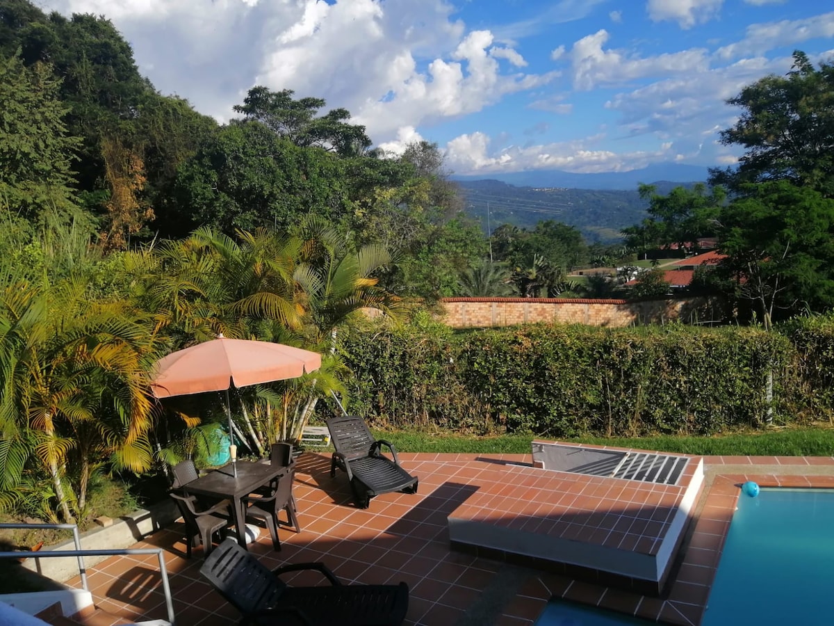 The outdoor area features a swimming pool surrounded by tiled flooring. A wooden table with chairs and an umbrella provide shaded seating. Lush greenery surrounds the space, enhancing the tranquil setting with views of distant mountains and blue skies.