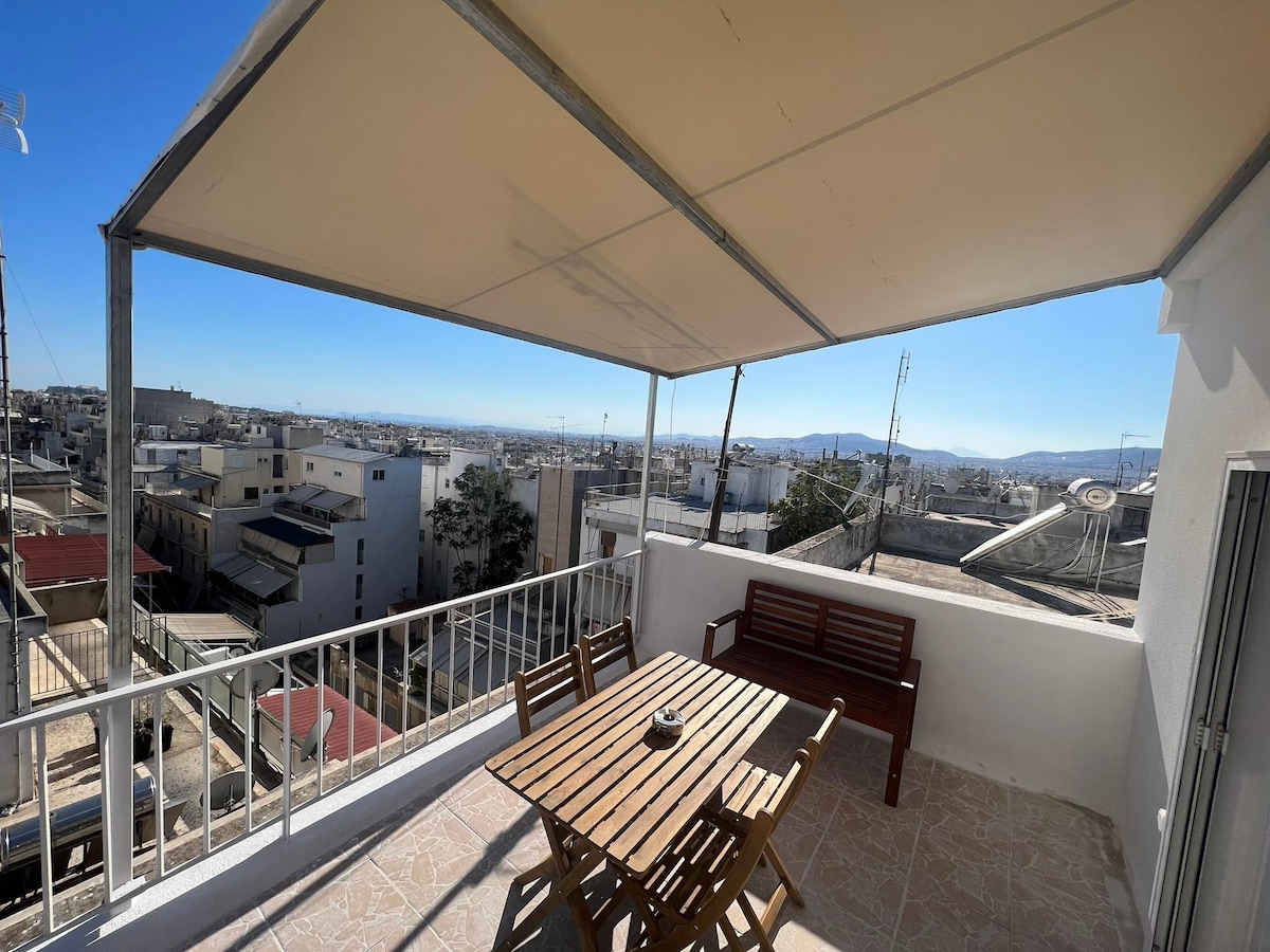 A private terrace is featured, offering a spacious area covered by a pergola. A wooden table with matching chairs is positioned for dining or relaxation, accompanied by a bench. The view reveals the cityscape and distant mountains under a clear blue sky.