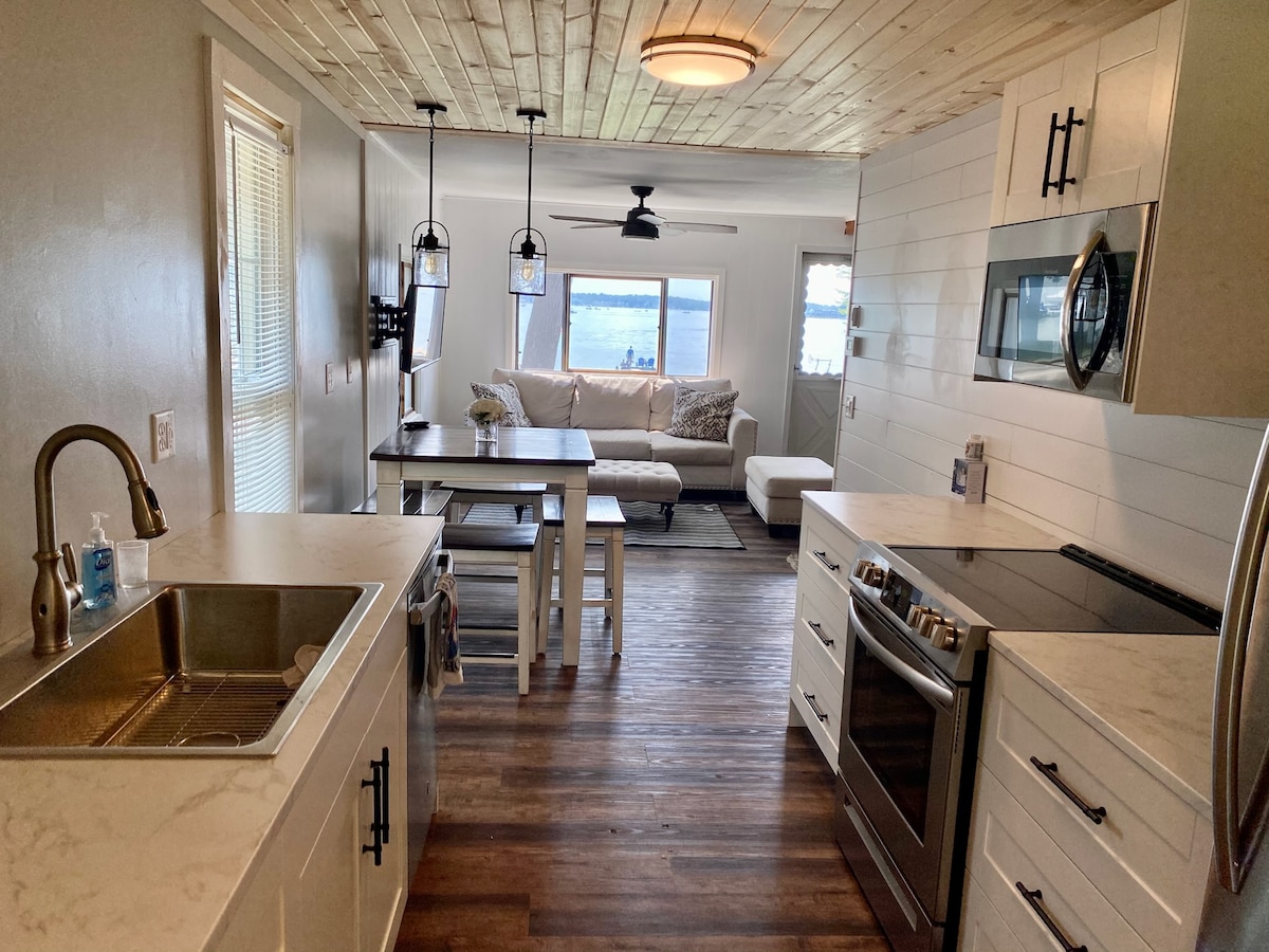 The kitchen showcases modern cabinetry and a sleek countertop, with a stainless steel sink and oven. A dining table with wooden seats is visible, along with a cozy living area featuring a light-colored couch and large windows overlooking the water.