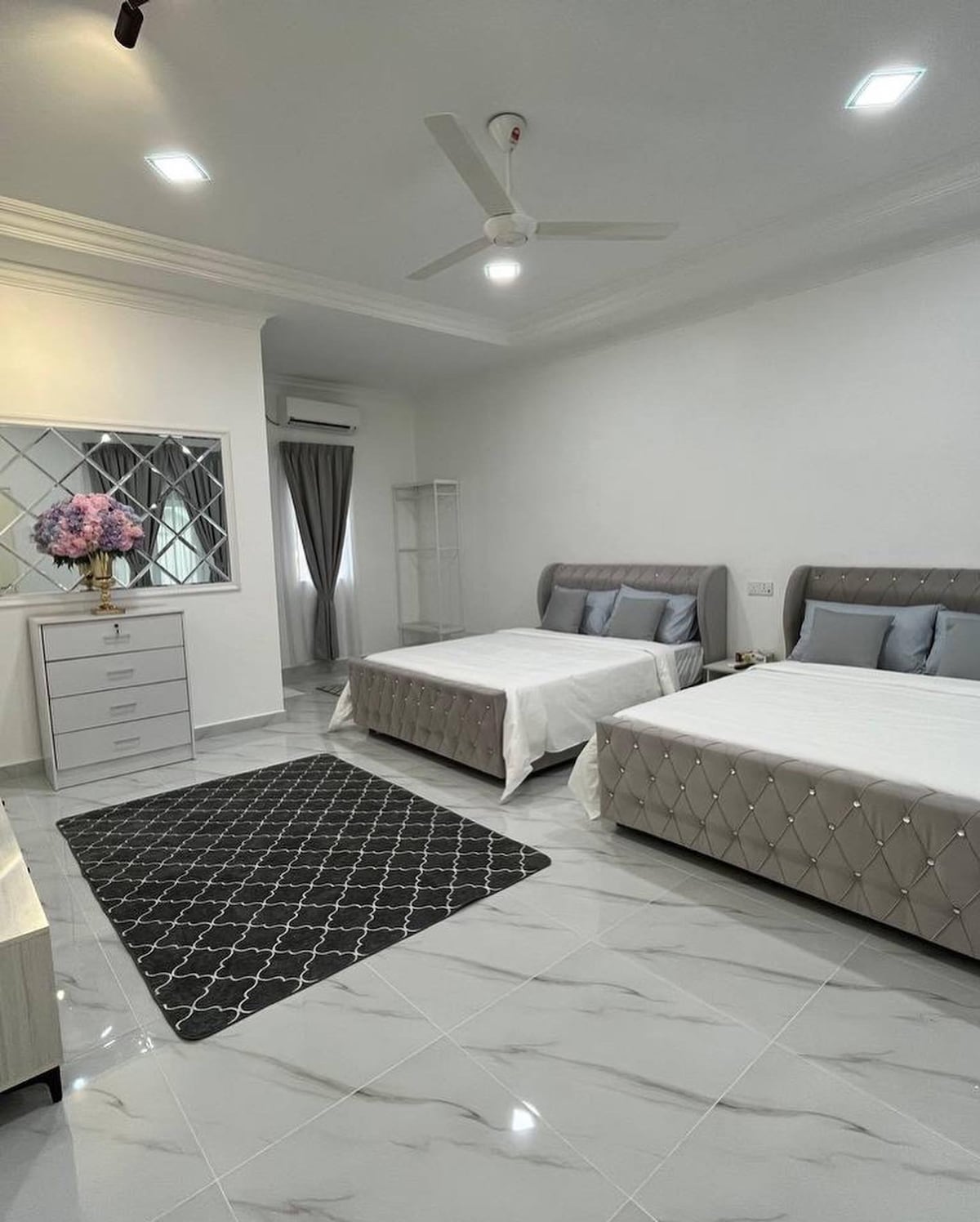 A bright studio room is furnished with two queen beds featuring upholstered headboards. A decorative area rug sits on the polished floor, and a contemporary dresser is positioned against the wall. Natural light is filtered through a window adorned with light curtains.