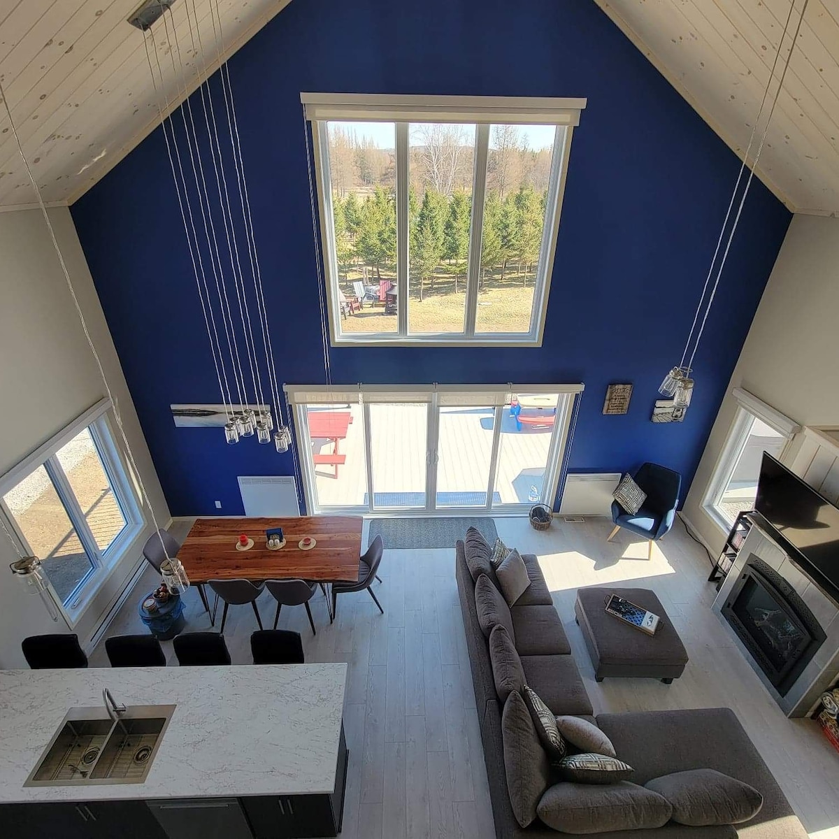 An aerial view showcases a spacious open-concept living area. A large sectional sofa and dining table are positioned near expansive glass doors, allowing natural light to fill the room. The blue accent wall adds depth, while views of the outdoor greenery are visible through the windows.