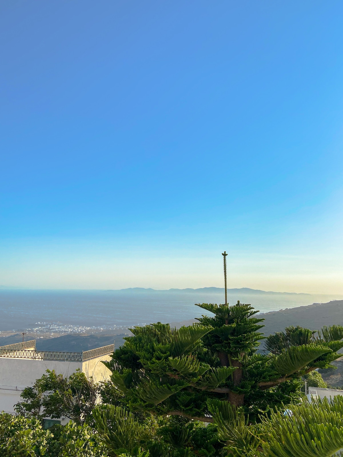 A panoramic view reveals the serene blue sky merging with distant islands and the vast sea. Lush greenery is featured in the foreground, enhancing the natural beauty of the landscape.
