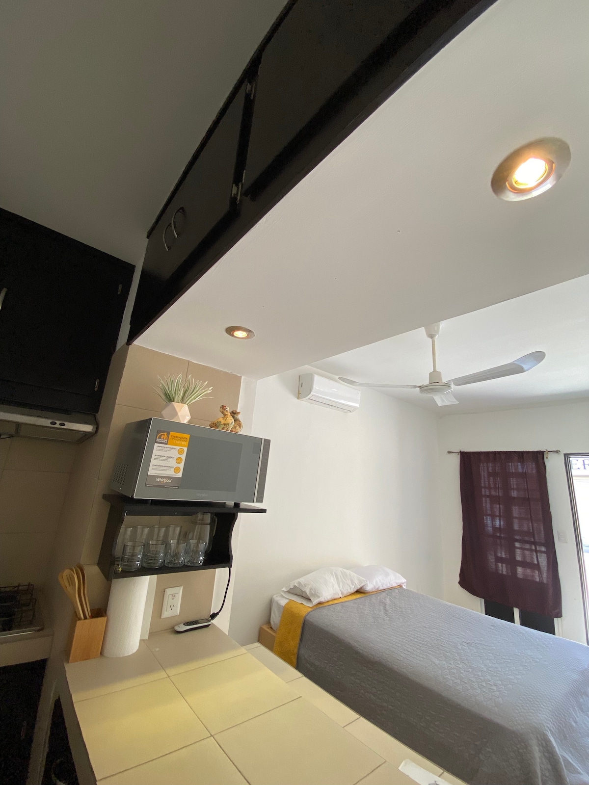 A compact living space features a light gray bed against the wall, complemented by a warm yellow accent. Above, cabinets provide storage, while a microwave and glassware are organized on the countertop. Soft lighting is provided by recessed lights and a ceiling fan.