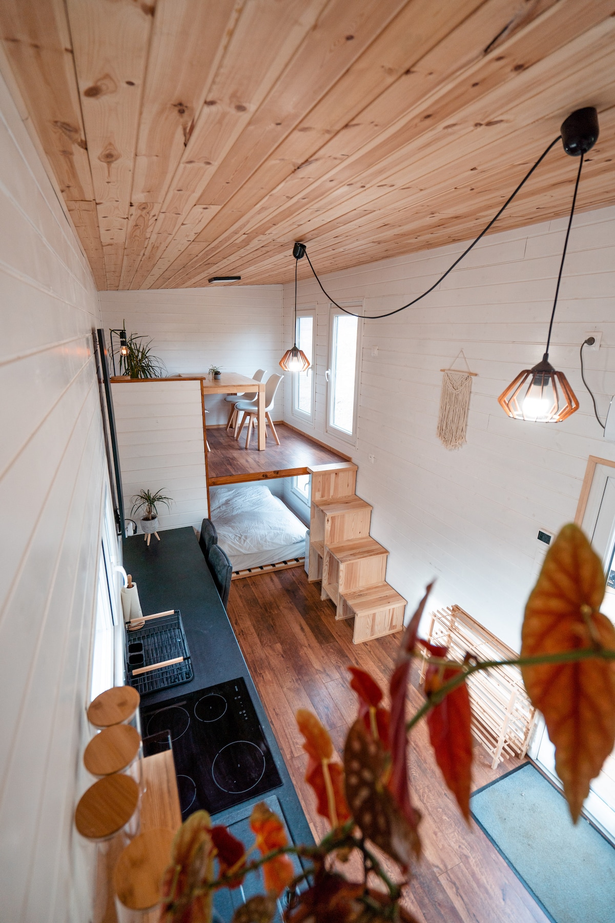 A well-lit interior features natural wooden accents and a high ceiling, showcasing an open living space. A wooden dining table is visible on a raised platform, while a staircase leads to an upper area. A cozy kitchen area is equipped with modern appliances and decorative plants.