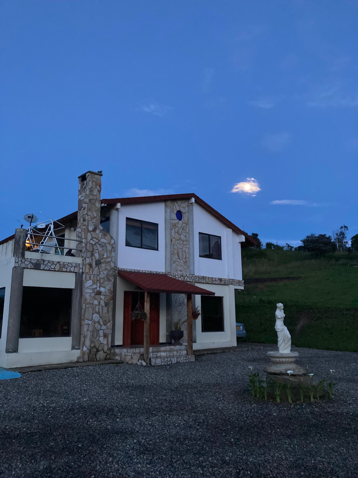 A two-story house features a stone facade and a red roof, set against a twilight sky. A small statue is positioned in the foreground, surrounded by gravel. The hills and distant greenery are visible, creating a serene backdrop.