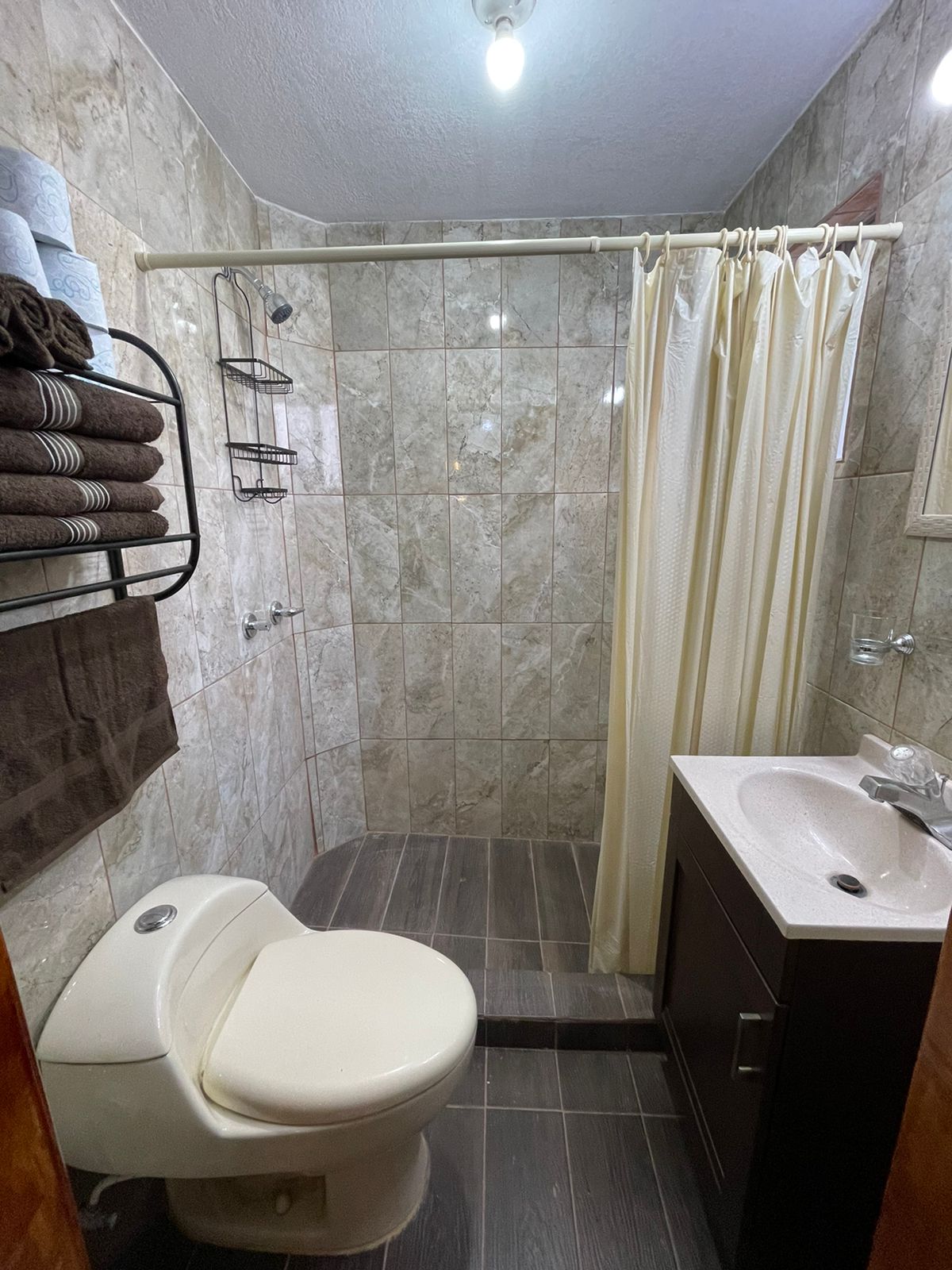 The bathroom features tiled walls with a neutral color scheme. A shower area is equipped with a curtain, while a toilet and a sink are present. Towels are neatly arranged on a wall-mounted rack, and additional storage shelves are provided for toiletries.