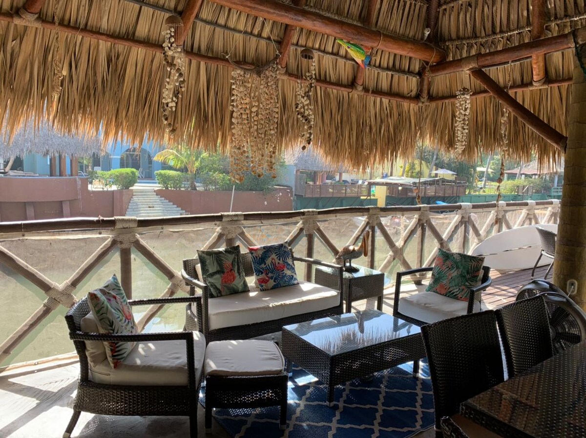 A shaded seating area is furnished with comfortable sofas and accent chairs, featuring decorative cushions. A low table sits in the center, surrounded by a natural straw roof. The serene canal views are visible through the railing, enhancing the outdoor experience.
