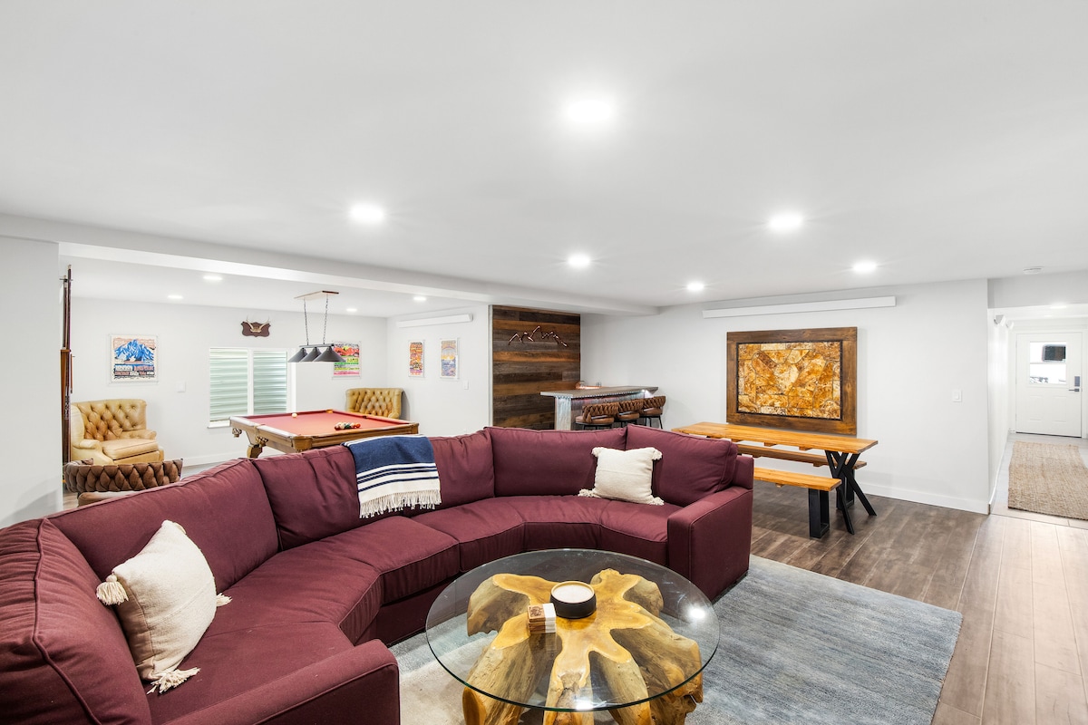 A spacious great room is visible, featuring a large sectional sofa surrounding a coffee table. An artistic piece hangs on the wall, and a dining table is set in the background. A pool table is positioned to the left, contributing to the inviting atmosphere.