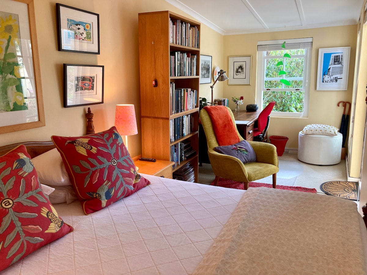 The spacious bedroom features a bed adorned with patterned pillows and a light-colored quilt. A wooden bookshelf filled with books, framed artworks, and a desk with a chair are visible. Soft lighting is provided by a lamp and natural light enters through a window with greenery outside.