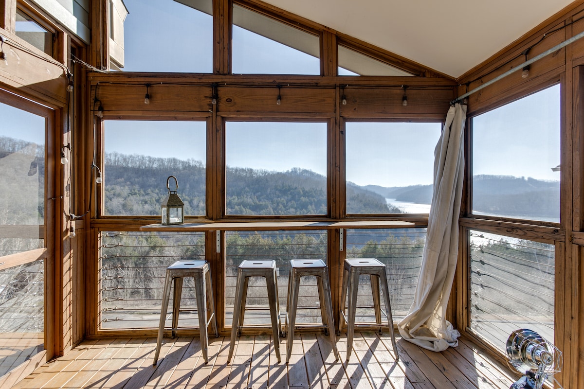 A bright, airy space is filled with natural light, featuring large windows that offer stunning views of the surrounding lake and hills. Four metal stools line the wooden deck area, creating an inviting spot to enjoy the scenery.