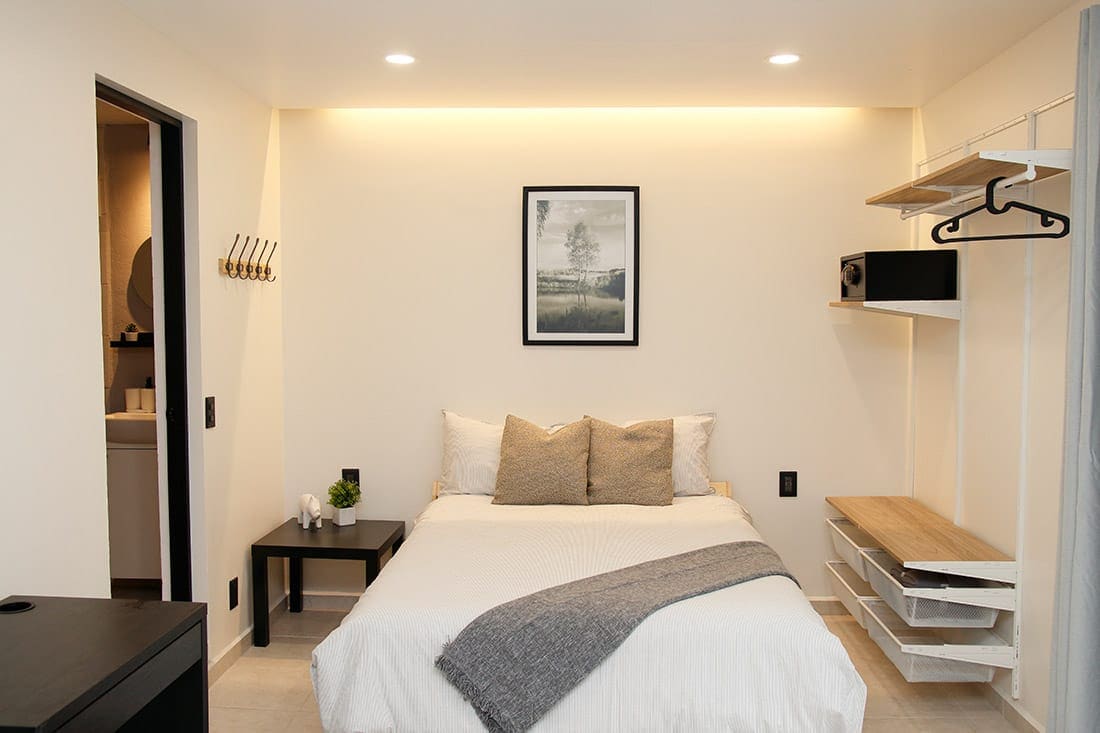 A welcoming bedroom features a double bed adorned with two decorative pillows. A framed landscape image hangs above the bed, while a small table sits beside it. Storage shelves and a hanging rod provide convenient space for belongings. Soft lighting creates a gentle ambiance.