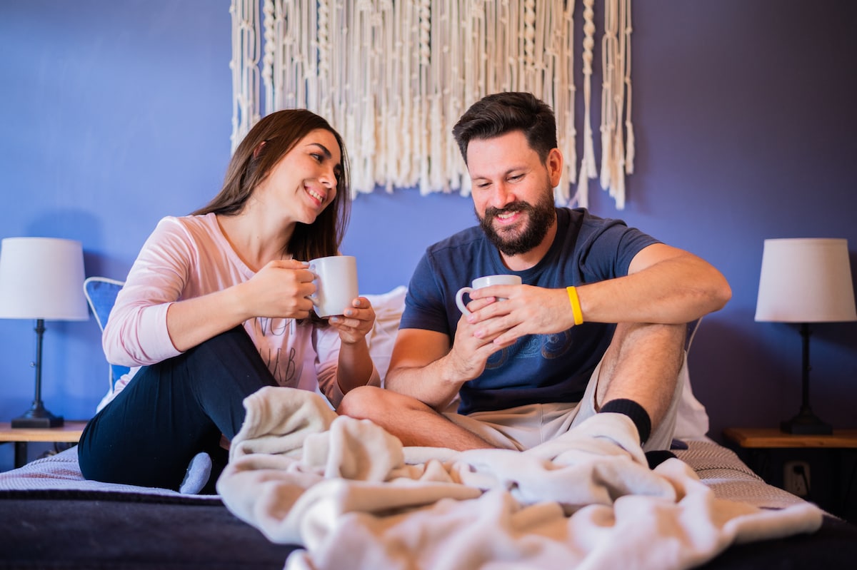 Two guests sit comfortably on a cozy bed, holding mugs. Soft blankets are draped across the bed. The backdrop features a textured wall hanging above the headboard, while warm bedside lamps create a relaxed atmosphere.