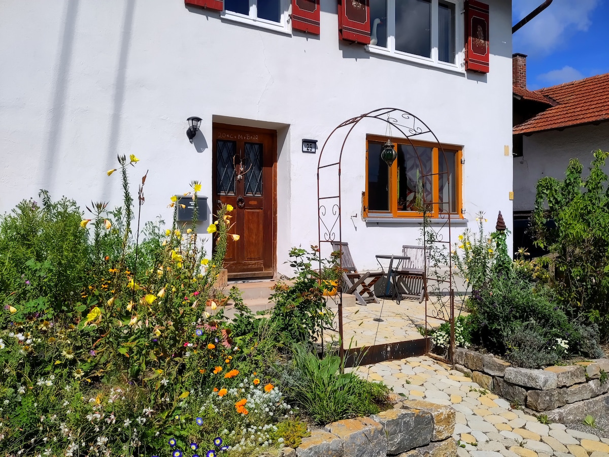 A charming entrance is framed by a blossoming garden filled with colorful flowers. An arched trellis leads to the wooden door, with two seating chairs set on a stone pathway. The white building features decorative shutters, enhancing the welcoming appearance.