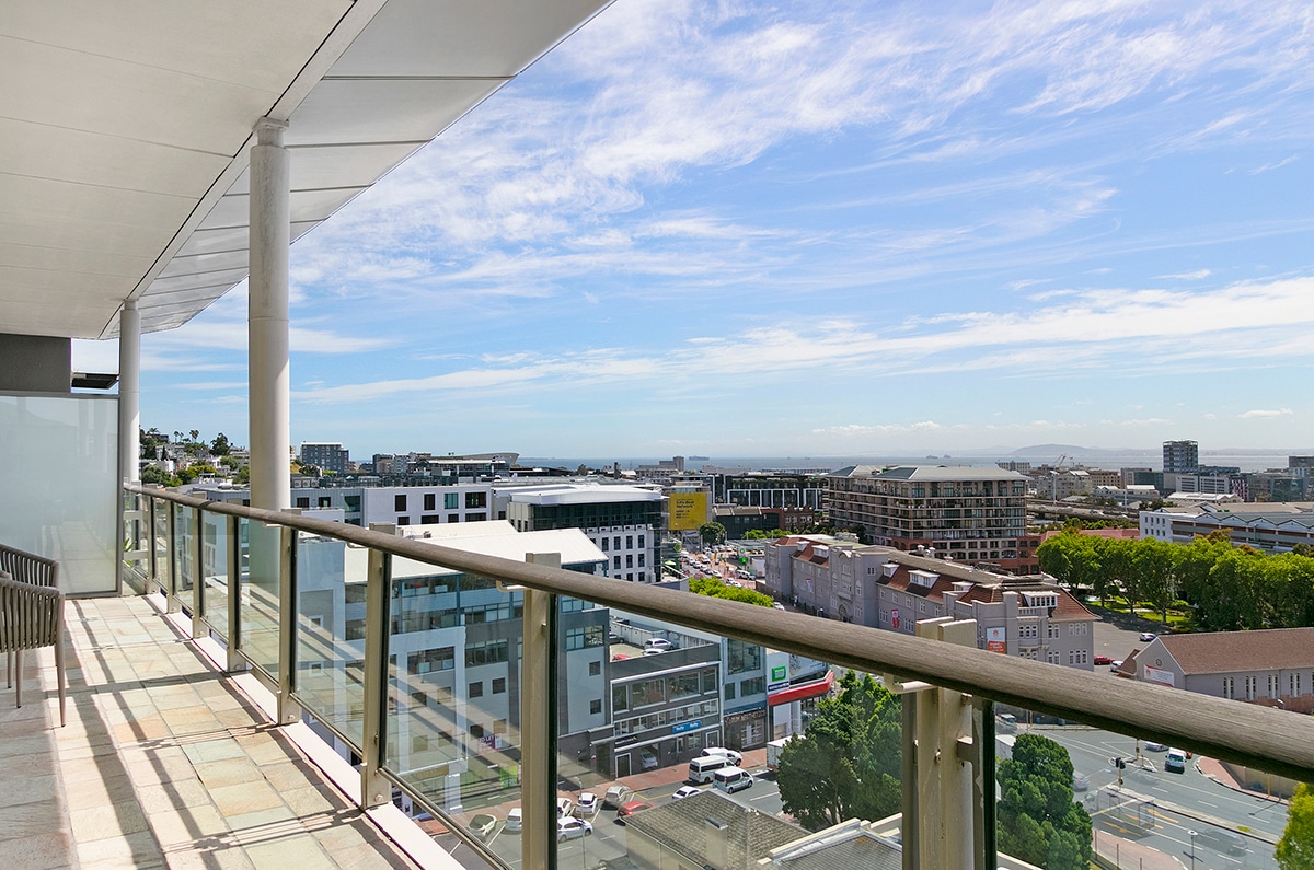 A spacious balcony is displayed, framed by glass railings that offer unobstructed views of Cape Town. The scenery includes distant ocean and urban landscapes, lit by natural sunlight. Two chairs are positioned on the balcony, inviting guests to enjoy the panoramic sights.