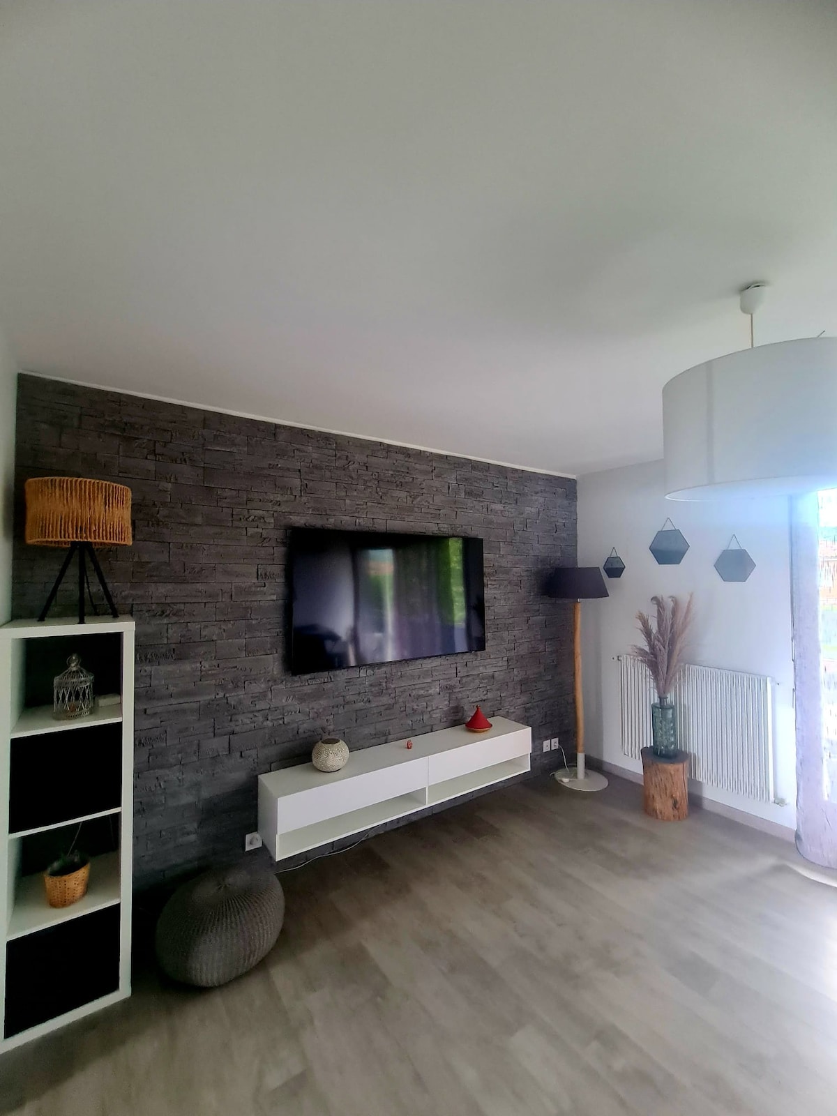 A living area features a sleek white media console below a mounted television. A textured dark stone accent wall adds depth, while a stylish pendant lamp and decorative items are placed on shelving units. Large windows bring in natural light, with a view of greenery.