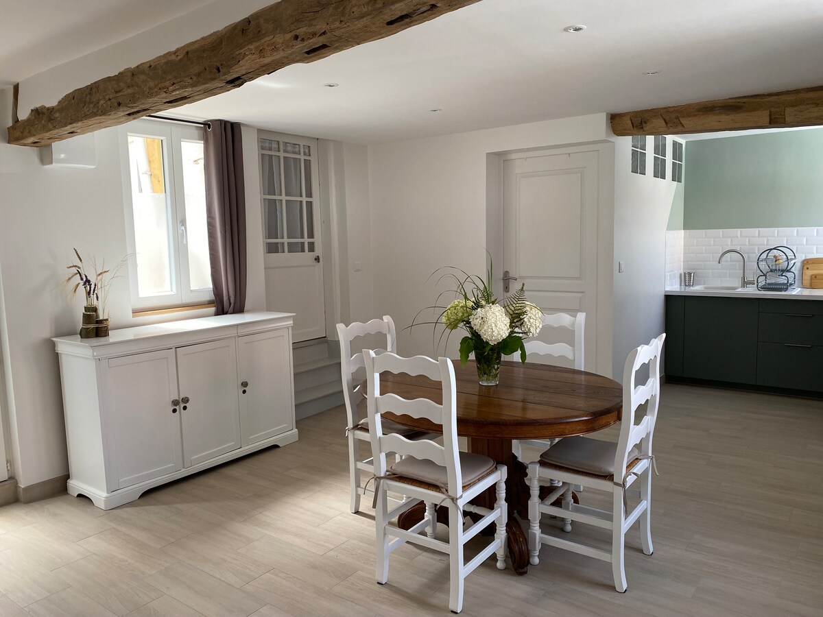 A spacious dining area features a wooden table complemented by six white chairs, creating a welcoming setting. Natural light enters through a nearby window, highlighting the room's soft tones. A sideboard with decorative elements rests against the wall, enhancing the inviting space.
