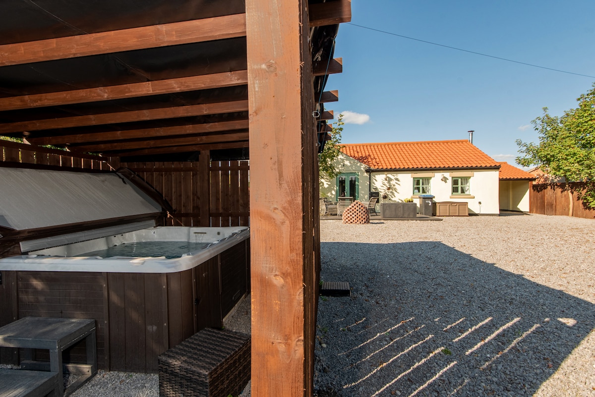 An outdoor area is showcased, featuring a well-maintained hot tub under a shaded structure. Gravel covers the ground, with a spacious layout that allows for various outdoor furniture arrangements. A cottage with green windows is visible in the background, complemented by clear blue skies.