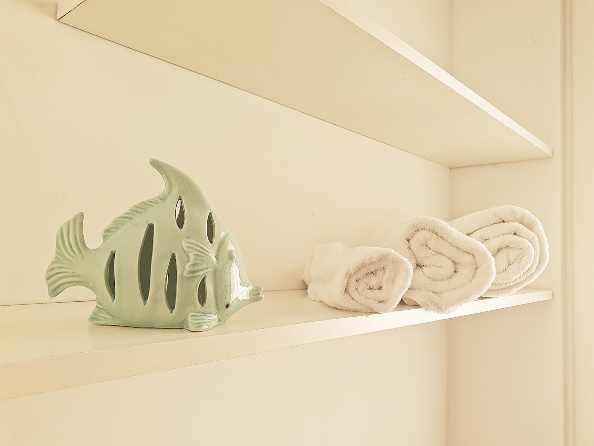A decorative ceramic fish in a soft mint color is placed on a white shelf next to neatly rolled white towels. The clean and simple arrangement highlights a touch of decor alongside essential towels.