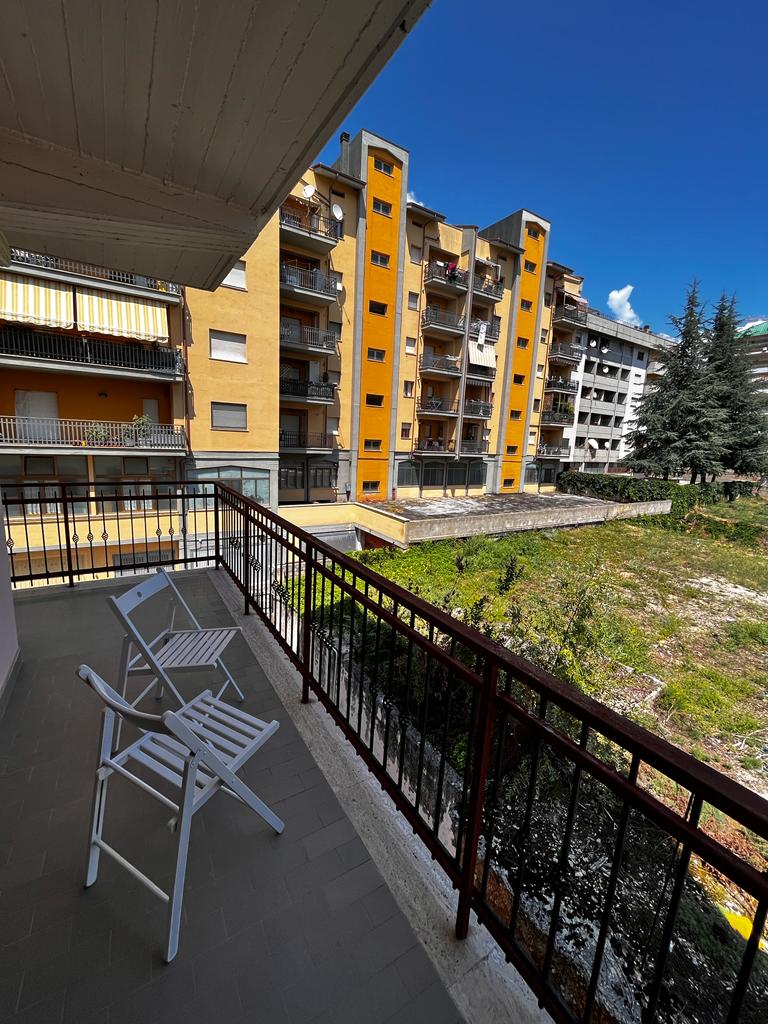A balcony is visible, featuring two white folding chairs with a small table. The view includes a sunlit area surrounded by greenery and several multi-story buildings, showcasing varied architectural styles and colors.