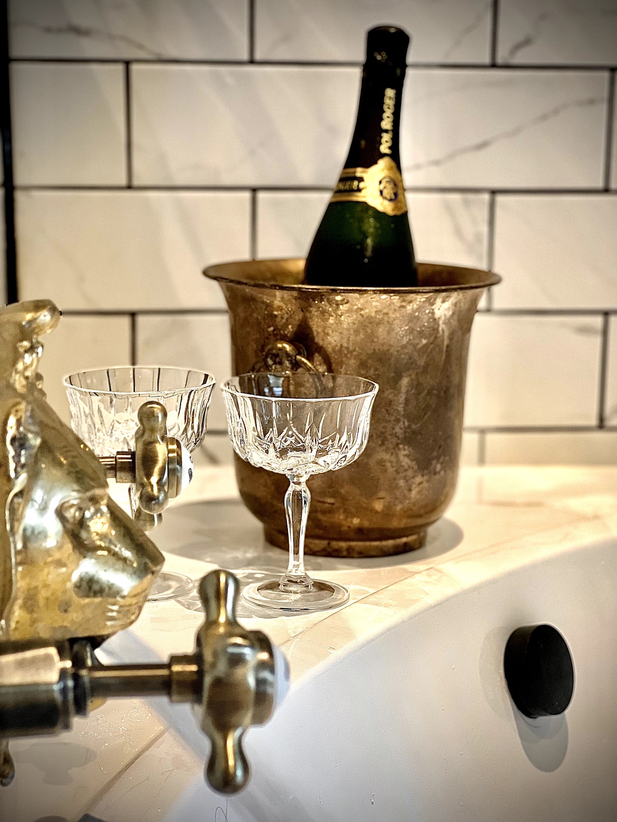 An elegant arrangement features a vintage champagne bucket filled with a bottle of sparkling wine. Two crystal glasses rest nearby, highlighting the luxurious atmosphere. The backdrop consists of stylish white tiles with subtle grey veining, enhancing the overall aesthetic of the setting.