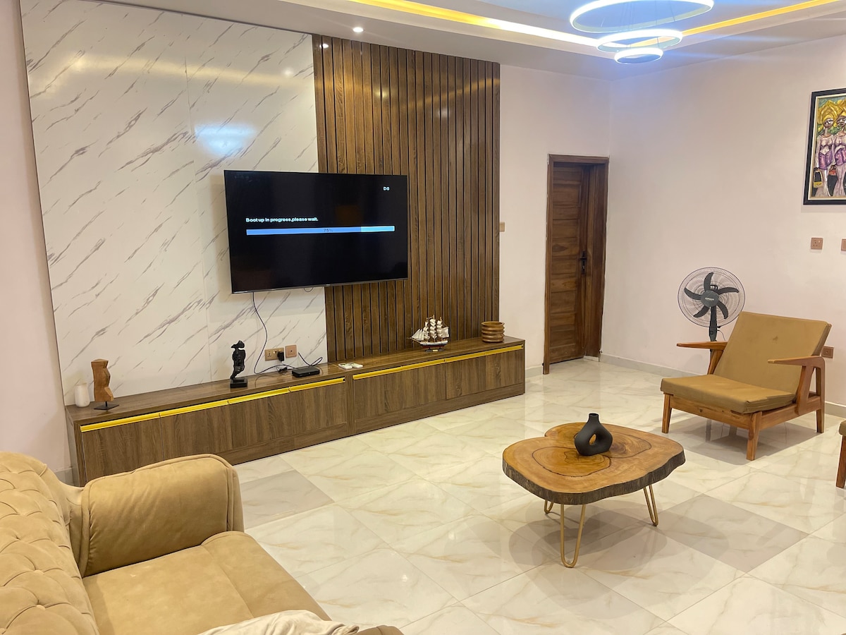 A modern living area features a large flat-screen television mounted on a sleek wooden wall. A light-colored sofa and a minimalist armchair complement a round coffee table. A ceiling light fixture creates a warm ambience, while a decorative fan adds functionality.