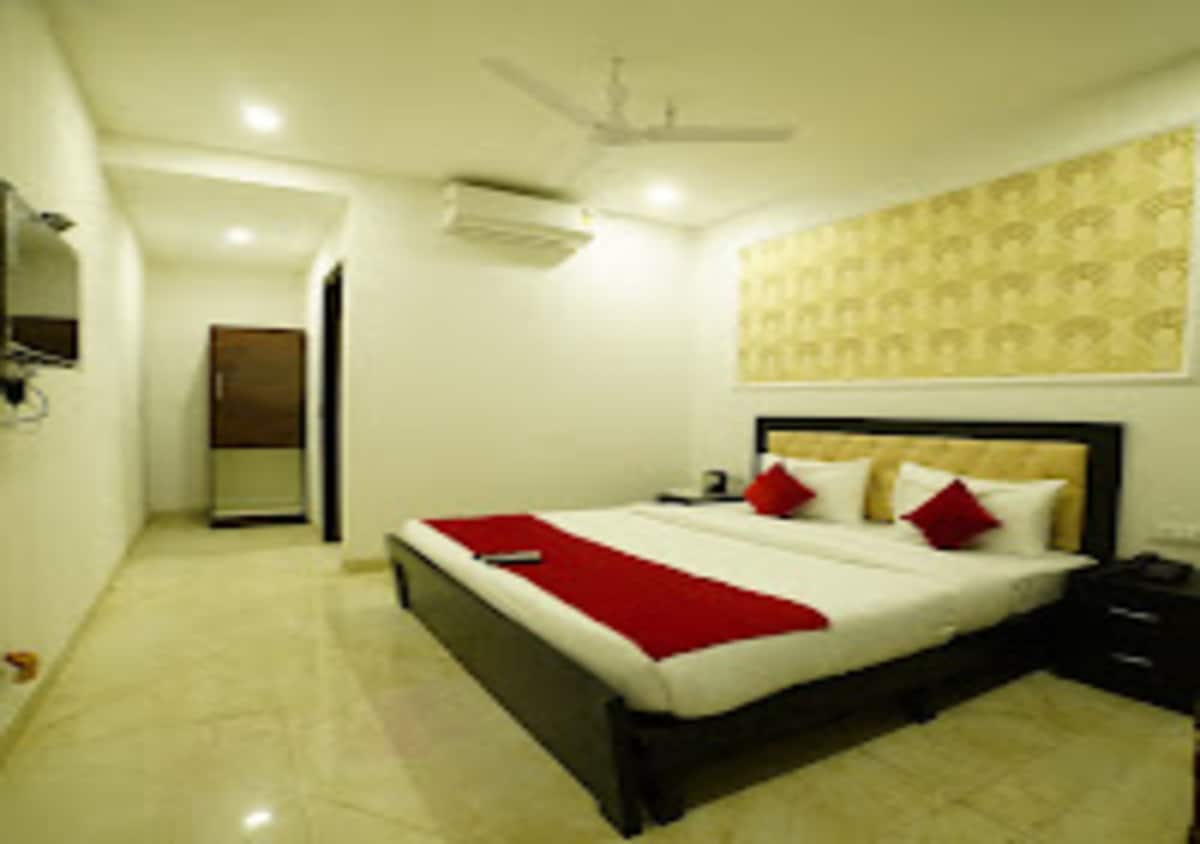 A bright bedroom is presented with light-colored walls and a tiled floor. A spacious bed is centered, adorned with white linens and red accent pillows. A television is mounted on the wall opposite the bed, and a closet is visible in the background.