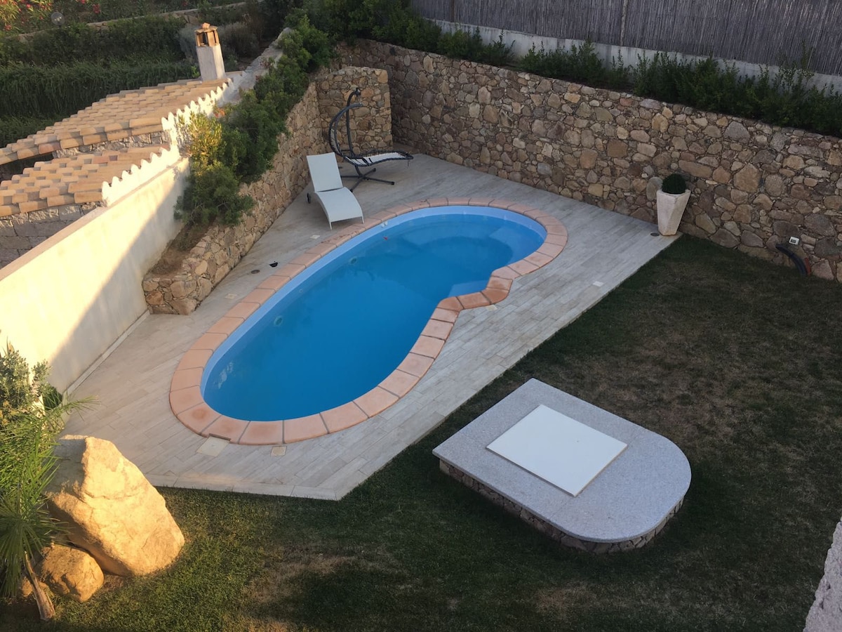 A serene outdoor space features an oval-shaped pool with a terracotta rim, surrounded by a stone wall. A sun lounger is positioned adjacent to the pool, while a rectangular table is set on a grassy area nearby, creating a relaxing environment.