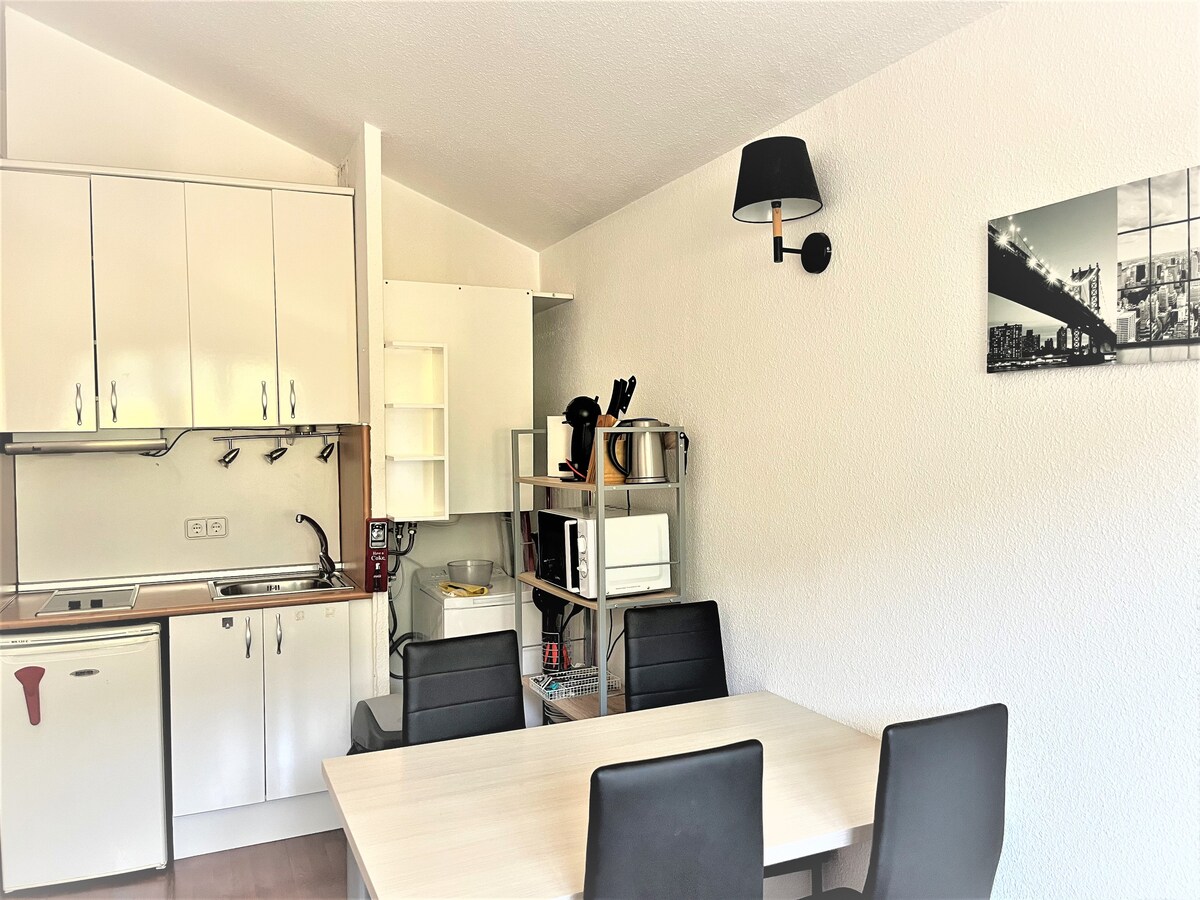 A well-equipped kitchen area is visible, featuring white cabinetry and essential appliances. A dining table for four black chairs stands in front of a light wall. A wall-mounted lamp provides ambient lighting, while a black and white photo adorns the adjacent wall.
