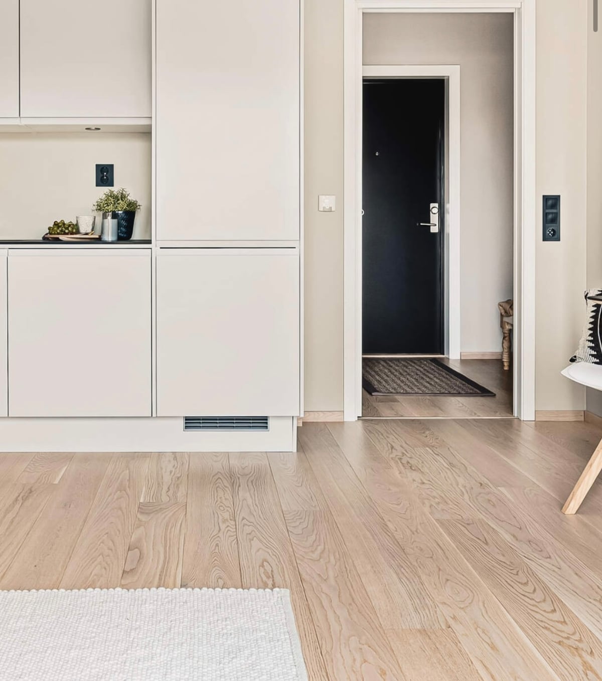 A clean and modern interior is displayed, featuring light wooden flooring and a minimalist kitchen area with sleek cabinetry. A doorway is visible, leading to an entrance with a dark door. A small rug and a chair are positioned in the foreground.