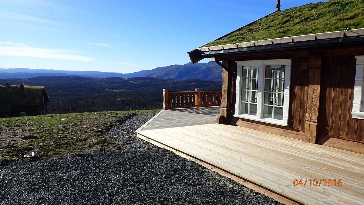 A large wooden deck extends from the cabin, offering sweeping views of the surrounding mountains and valleys. The cabin features multiple windows that allow natural light to fill the interior. The grassy roof blends harmoniously with the landscape.